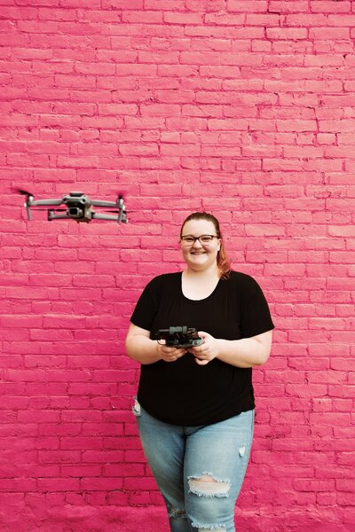 mara in front of a pink brick wall, flying a drone with the drone slightly in front of her closer to the camera