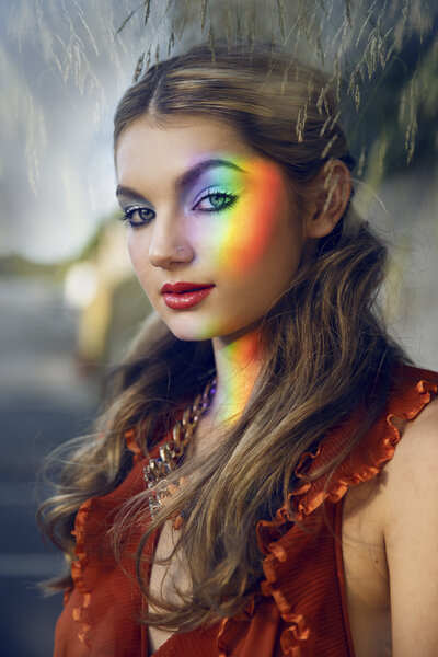 Magical outdoor senior portrait of a girl with glowy makeup and a rainbow reflection cast across her face. Her colorful eyeshadow and soft curls complete this dreamy, whimsical look.