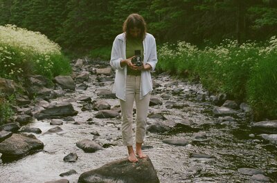 Heather stands on a rock in the middle of a river, taking a photo on a TLR film camera. They are a white person with brown hair, wearing all white.