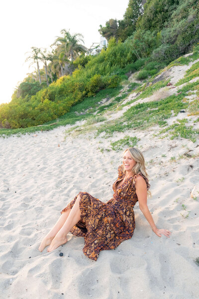 A woman life coach sitting on the sand at the beach wearing a dress feeling mental clarify and freedom.