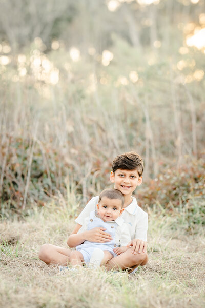 The picture captures a heartwarming moment between two brothers in a serene, wintry setting. The older brother, dressed smartly in a button-down shirt and slacks, tenderly holds his baby sibling in his lap. The younger boy rests comfortably against his big brother's chest, their bond palpable in the gentle way the older child cradles him.  The scene is set against a backdrop of a lush, forested area, the bare trees and dry grass hinting at the winter season in Texas. The lighting is soft and airy, casting a warm, ethereal glow over the brothers, highlighting the delicate features of their faces and the love between them.  The photographer, Bri Sullivan, has masterfully captured the essence of this sibling relationship, the older brother's protective instinct and the younger's complete trust in him. The composition is well-balanced, with the brothers occupying the central focus, while the natural surroundings provide a beautiful, understated setting.  Overall, this photograph is a stunning testament to the power of familial love and the special connection between siblings. It is a moment frozen in time, a glimpse into the tender, intimate relationship between these two boys, and a testament to the skill and artistry of the photographer who captured it.