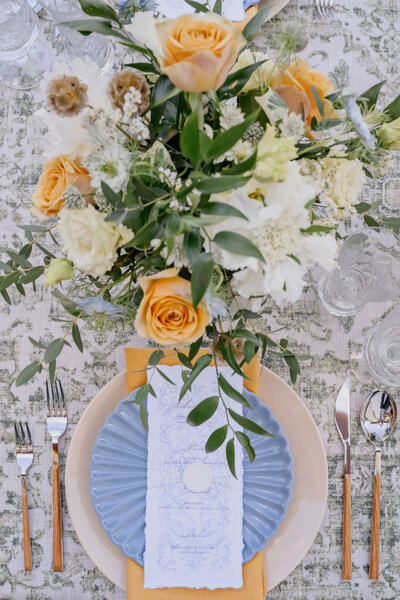 A top-down view of a luxurious tablescape featuring a vibrant floral centerpiece with yellow roses, blue scalloped plates, handwritten menus, and gold-accented flatware