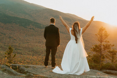 Bride and groom celebrating their marriage on the edge of a cliff in the White Mountains of New Hampshire as the sun sets and golden light fills the frame.