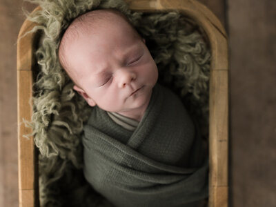 newborn baby boy wrapped in green swaddle laying in basket for studio newborn portraits
