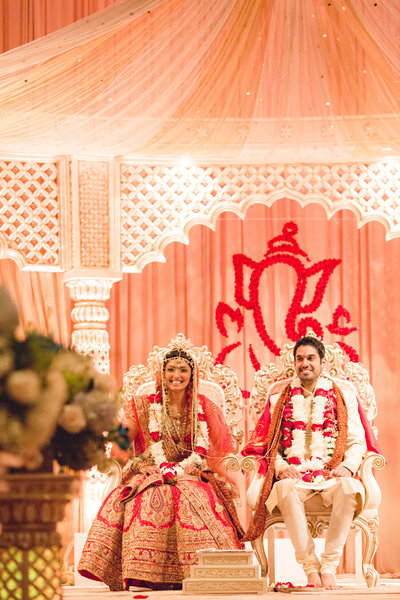 A bride and groom in traditional Indian wedding attire sitting under an ornate canopy with a decorative backdrop, planned by a top Illinois wedding planner.