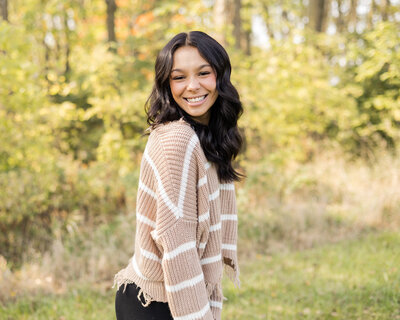 high school female senior, with long black hair, standing in grass with a tree line next to her, standing to the side, looking at the camera smiling, wearing black leggings, and light tan and white stripped shirt