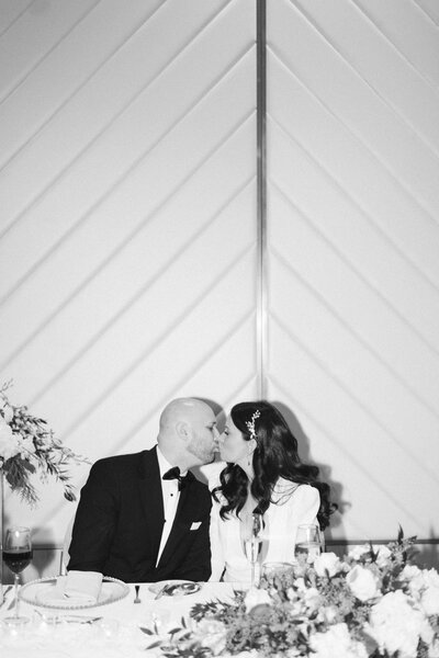 Burlington, Ontario Bride and groom sharing a kiss at their reception head table, surrounded by florals at The Pearl Hotel.