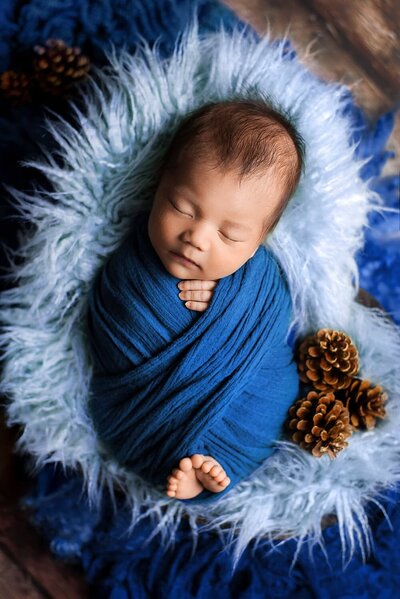 Newborn baby boy wrapped in a criss-cross style blue wrap, posed in a heart basket during an in-home session.