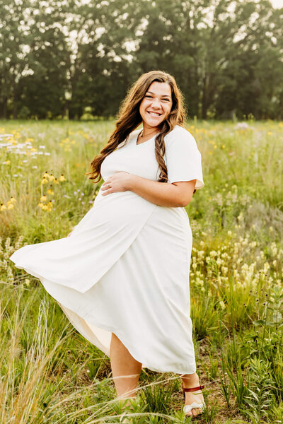 Pretty mom to be in a white dress resting hand on her baby bump as she dances in a flower field near Green Bay