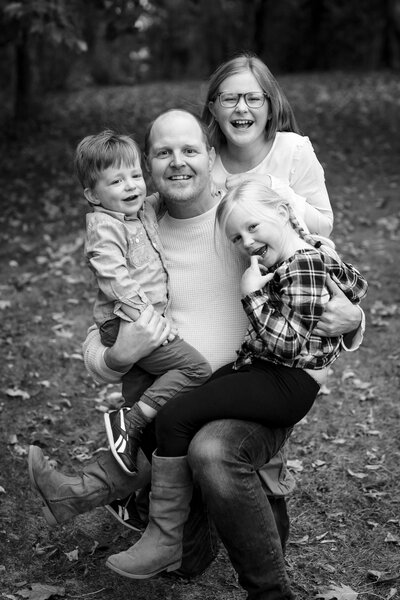 Black-and-white family portrait at Patterson Park in Kingston, Ontario, featuring a father kneeling and holding his two younger children, with his oldest daughter standing behind him smiling warmly.