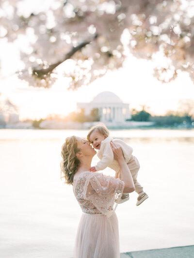 A cherry blossom photo session at the Tidal Basin with a mother holding up her young son with the Jefferson Memorial in the distance.
