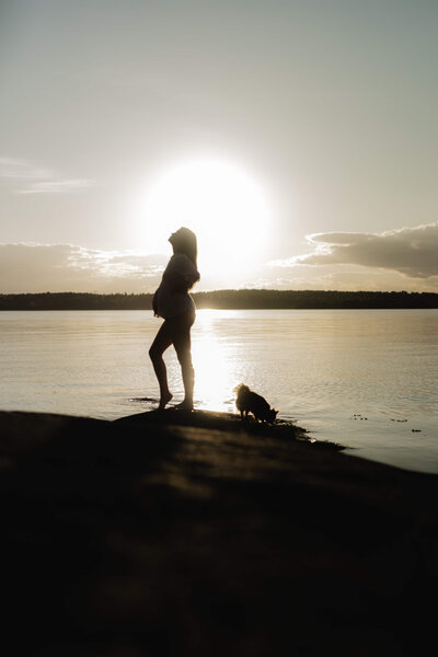 En gravid kvinna ståendes ute vid vattnet tillsammans med sin hund under solnedgången under en gravidfotografering i Stockholm