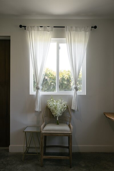 Minimalist interior scene with a bouquet of white orchids resting on a woven chair beneath sheer white curtains and a sunlit window — captured in a natural light-filled room in Vero Beach, Florida.