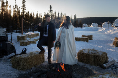 Bride and groom stand warming up by a bonfire on their snowy winter elopement day. The groom holds an axe and has his foot up on a straw bale, as the bride laughs.