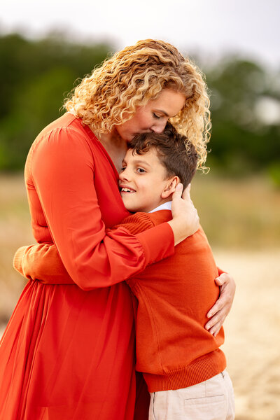 A mother embraces her son in a heartfelt hug during a fall family session, both wearing matching red outfits as golden light pours through the trees in Virginia Beach
