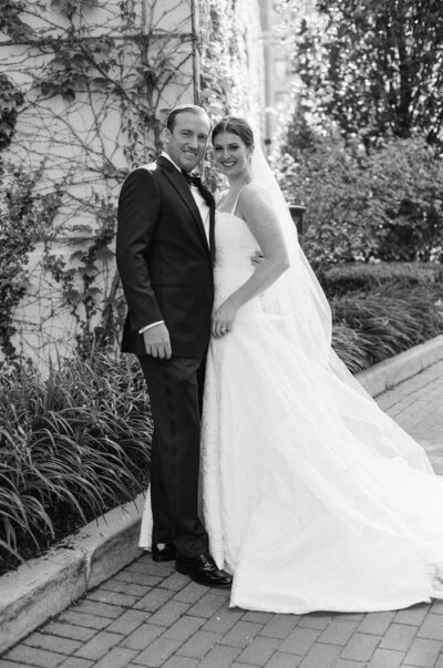 A bride in a flowing white gown and a groom in a classic black suit share a joyous moment together, posing in a lush garden setting captured in a timeless black and white photo.