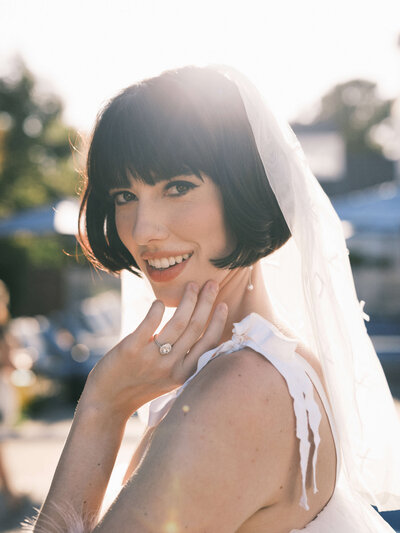 A fun bridal portrait of a bride with a short bob smiling right into the camera  while the sun shines through their veil