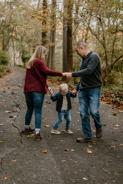 Picture of mom, dad, and toddler boy in a wooded park playing ring around the rosie in the fall in Anne Arundel County Maryland