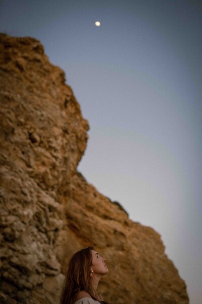 A woman with long hair looks upward at the glowing moon in the early evening sky, standing beside rugged golden cliffs under soft twilight.