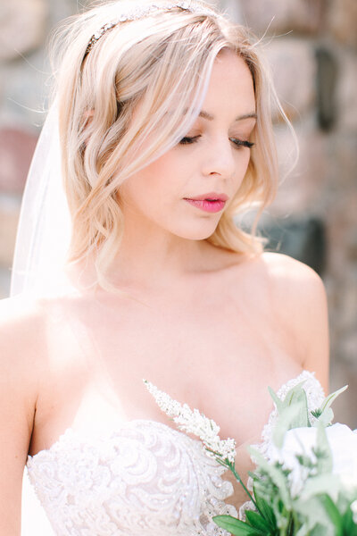 A blond brides admires her floral bouquet. She wears a soft pink lip with flushed cheeks.