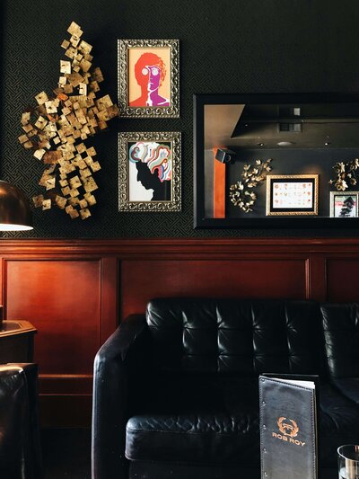 A cozy lounge with dark walls, modern artwork in ornate frames, and a black leather sofa against a polished wood panel backdrop. A menu labeled "Rob Roy" sits on a table in the foreground.