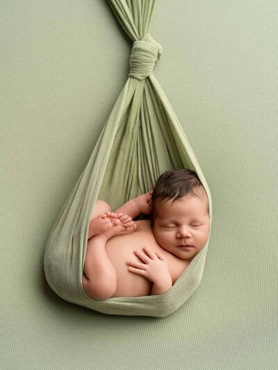 A peaceful baby sleeps curled up in a soft green hammock against a matching textured background, conveying comfort and serenity.