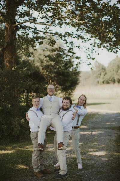 Three groomsmen in white shirts and suspenders lift the groom, who is wearing a beige vest and pants, as they all laugh together on a sunny outdoor path surrounded by trees.
