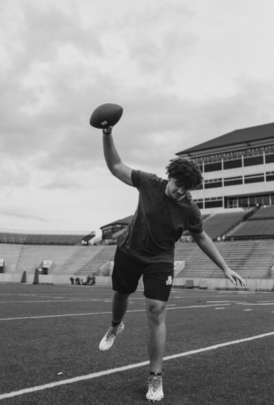 Black and white image of a male athlete holding a football in his right hand like he's going to throw it down in a stadium