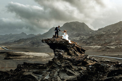 bride and groom are climbing up on a cliff and surrounded by mountains in Iceland