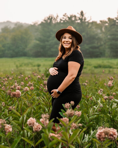 pregnant mom shows off her bump in tight black dress and brown hat in a field of flowers