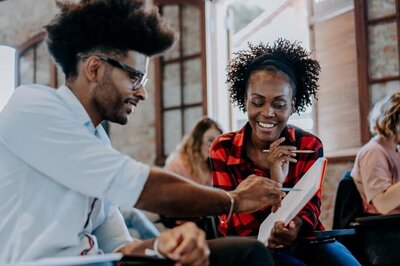 Two people in a casual workshop setting smile over a notebook. Left, a person with an afro and glasses points to the page; right, a person in a red plaid shirt and headband holds the notebook and a pen, engaged in the discussion.