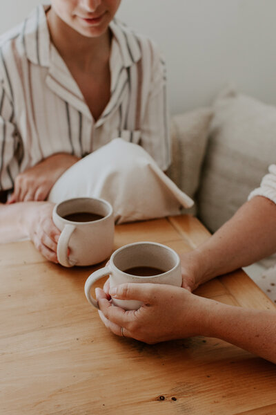 2 women sitting at a table drinking mugs of coffee 