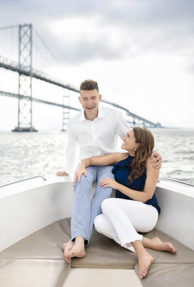 A couple sits on a boat, enjoying a sunny day. The man, wearing a white shirt and light blue pants, smiles at the camera. The woman, in a navy top and white pants, gazes up at him. A large bridge stretches across the background.