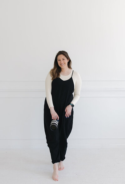 Tia Quirk in a black jumpsuit on a white background holding a camera, ready to capture wedding photos of local couples in MN