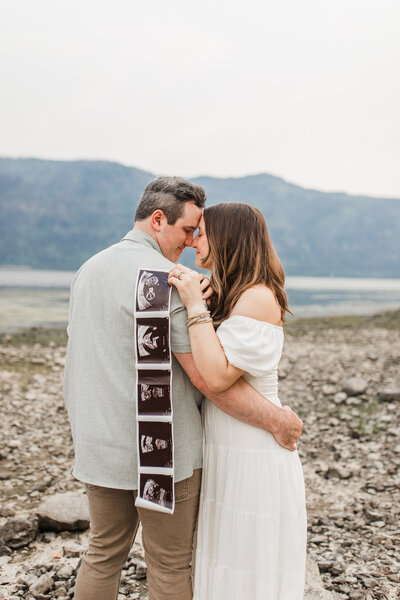 Portland Newborn Photographer_ pregnancy announcement photos featuring a couple smiling forehead to forehead while holding ultrasound photos and the beautiful Columbia river and blue mountains in the backdrop