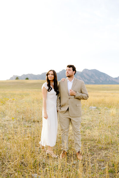 Engaged couple Annie and Jared standing in a golden field with the Flatirons mountains behind them during their Colorado engagement session, captured at sunset in elegant neutral outfits.