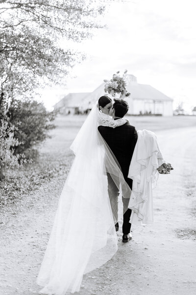 Black and white photo of groom carrying bride with venue in the background