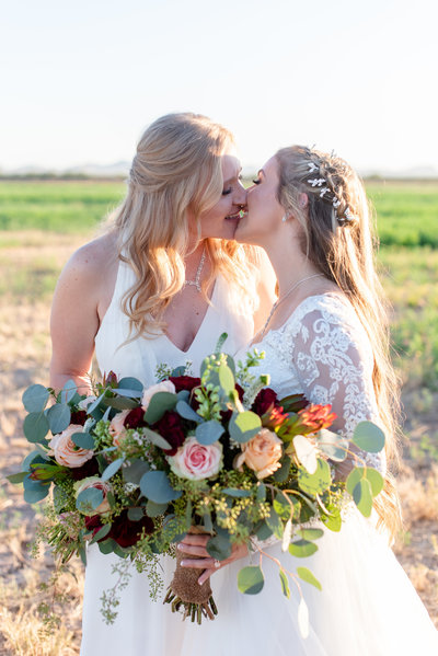 Two brides go in for a kiss holding flowers in a field at Wellington Ranch