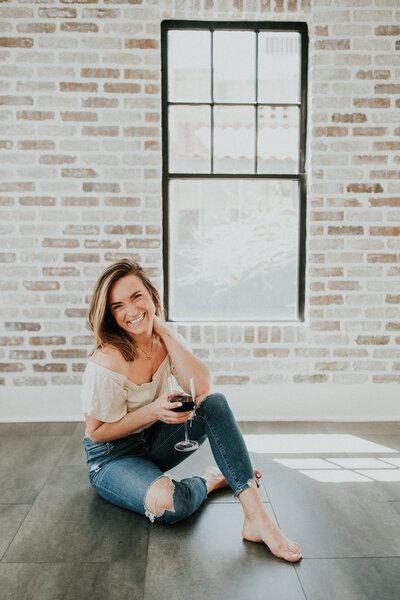 Missoula Montana photographer sits on the floor as she holds a wine glass in front a brick wall.