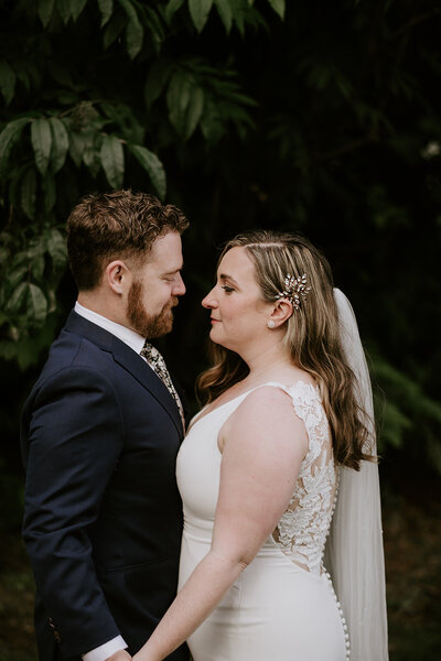 A bride in a white gown and a groom in a navy suit face each other outdoors.