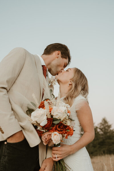 A photo taken by Bailee Freeman Photography of a bride and groom kissing. Photo is taken at a low angle so the sky is the main background and the bouquet is at the bottom of the photo. 