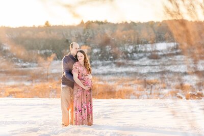 Couple stands in snow during winter maternity photo session with Sara