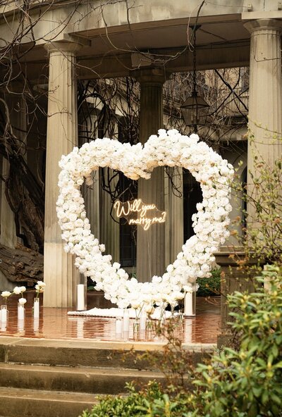 Romantic proposal backdrop with a large white floral heart arch and ‘Will you marry me?’ neon sign, styled by Yours Truly, a wedding planner and designer in New Jersey.