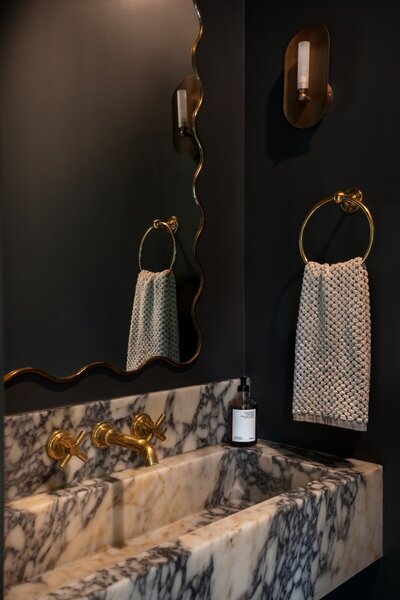 Guest powder room with black walls, wavy gold mirror frame, gold hardware, and black and grey marble countertop.