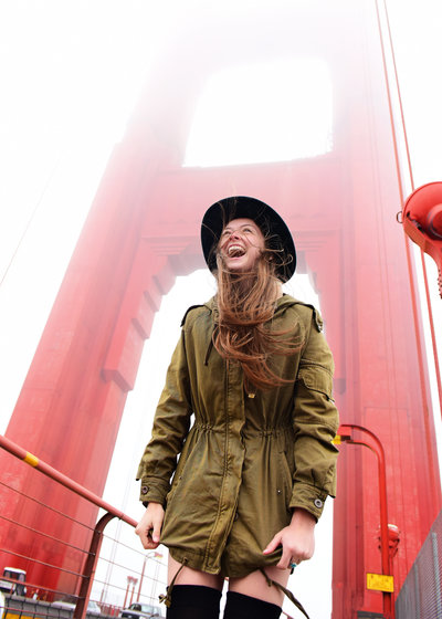 Adventurous young lady wearing a green coat thigh black socks black Australian  hat laughing as she stands on the San Francisco Bridge that is bright red