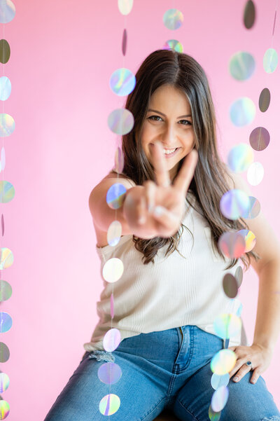 Bee, owner of BusyBSocial, puts up the peace sign at the camera while surrounded by draping holographic streamers, her other hand is on her hip playfully and she smiles in front of a pink backdrop