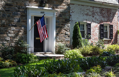 Beautifully landscaped front yard of a historic stone and brick home, featuring neatly trimmed plants, a lush green lawn, and a refreshed entrance with an American flag.