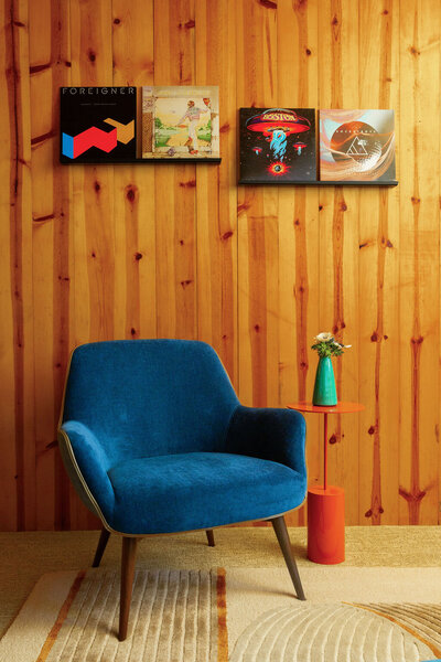 A cozy corner with a vibrant blue Gretchen chair on a patterned rug against a wood-paneled wall. Above the chair, three vinyl record albums in black frames are mounted on the wall: "Foreigner," an album with a landscape scene, and "Boston." A small orange cylindrical side table next to the chair holds a teal vase with white flowers.