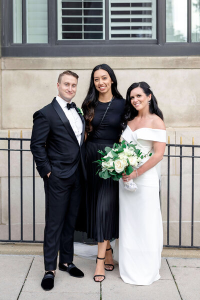 A groom in a black tuxedo and a bride in an off-the-shoulder white dress stand on either side of stellar wedding planner Lisseth Orellana. The bride holds a bouquet of white roses. They pose in front of a building with iron fences.