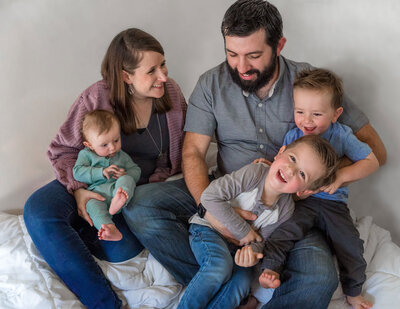 A family with three boys under 4, dressed in pastel colors, laughs and interacts with each other as they sit together on a blanket in front of a white background. Photo by SAVI Photography - Photographer in Riverside